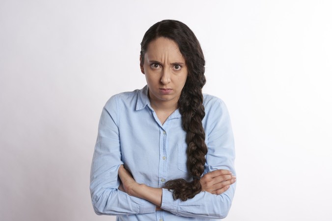 A white woman with long, curly, dark brown hair in a plait. She glares at the viewer with arms tightly crossed. She is wearing a pale blue blouse.