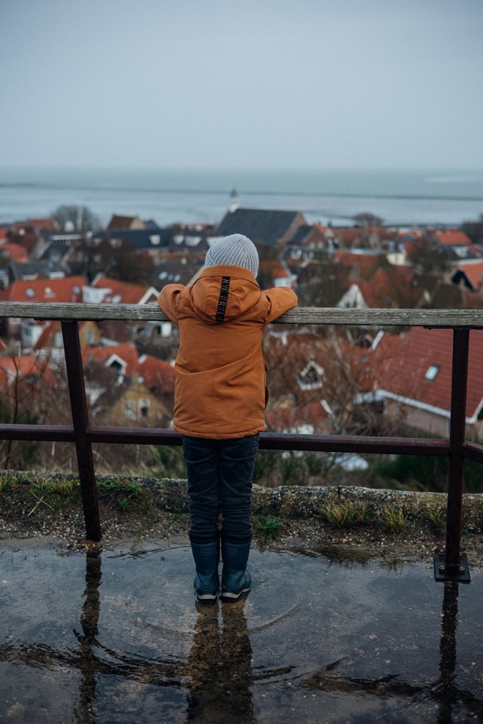 A boy wearing an orange coat leans on a railing overlooking a coastal town.