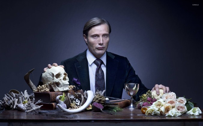 Picture of actor Mads Mikkelsen portraying Dr. Hannibal Lecter. He sits at a table with one hand on a human skull and the other on a cauliflower. There is a silver wine goblet on the table as well as several leather bound books, various bones, vegetables, and white roses.